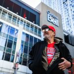 Older man in a red cap and Canadiens shirt stands on a busy city street in front of a glass-fronted building with a Bell sign (urban scene).