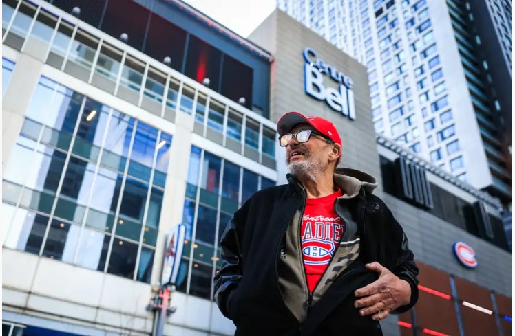 Older man in a red cap and Canadiens shirt stands on a busy city street in front of a glass-fronted building with a Bell sign (urban scene).