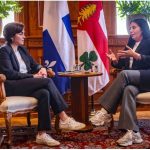 Two women in formal business attire sit facing each other in ornate armchairs, with flags displayed behind them during a meeting.