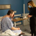 Patient in a hospital gown sits in a chair while a clinician wearing a mask checks him with a stethoscope nearby at a bedside table stay.