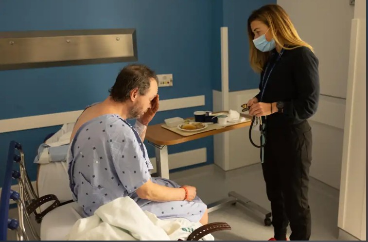 Patient in a hospital gown sits in a chair while a clinician wearing a mask checks him with a stethoscope nearby at a bedside table stay.