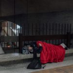 Person sleeping on a concrete bench under a bridge, covered with a red blanket and a backpack nearby (street scene)