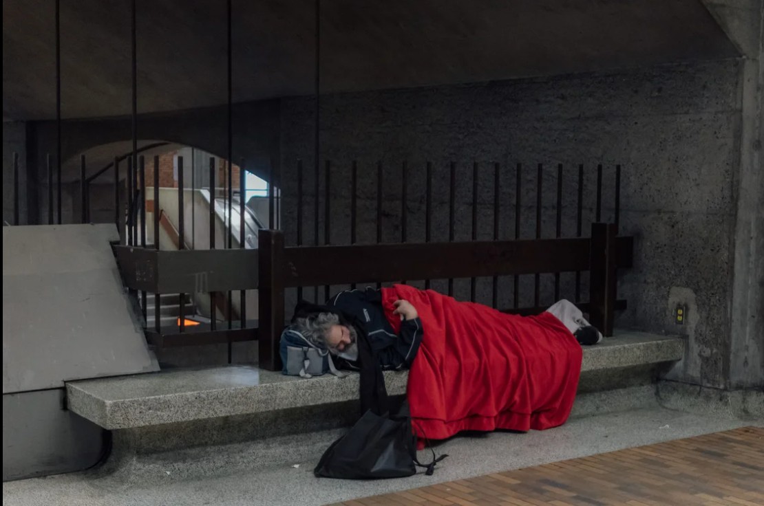 Person sleeping on a concrete bench under a bridge, covered with a red blanket and a backpack nearby (street scene)