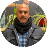 Portrait of a bald man wearing a black scarf and denim jacket, looking at the camera with plants in the background.