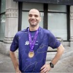 Smiling man in a purple running shirt with a medal around his neck, standing on a sidewalk in front of a storefront.