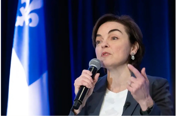 Woman in a blazer speaks into a handheld microphone on stage, with a blue flag in the background.