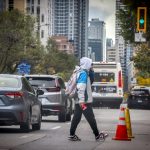Pedestrian in a hoodie and backpack crossing a city street with a bus approaching behind and traffic cones nearby.