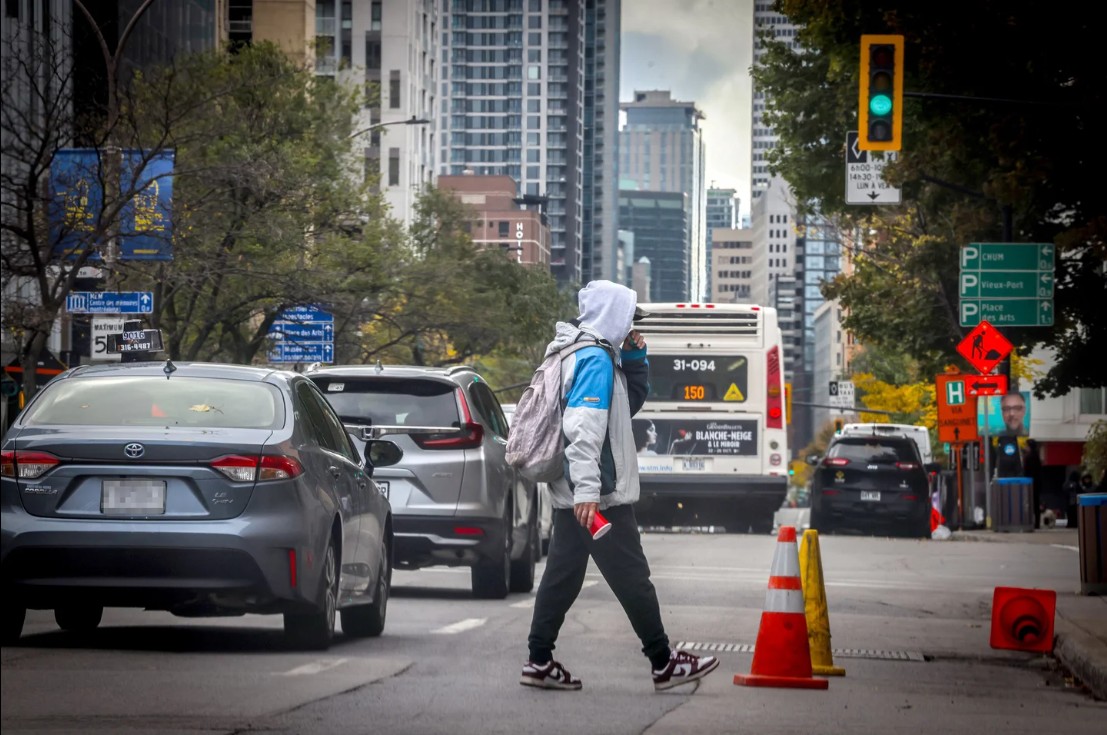 Pedestrian in a hoodie and backpack crossing a city street with a bus approaching behind and traffic cones nearby.