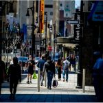 Busy city sidewalk with pedestrians, storefronts, and street signs; some people wearing masks.