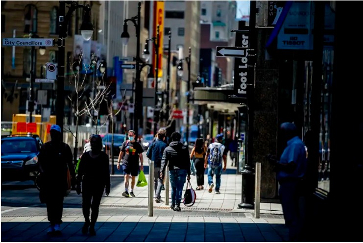 Busy city sidewalk with pedestrians, storefronts, and street signs; some people wearing masks.