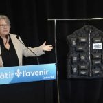 Woman in a beige blazer speaks at a podium labeled 'Bâtir l'avenir Québec' with a black tactical vest on a stand beside her, during a presentation.