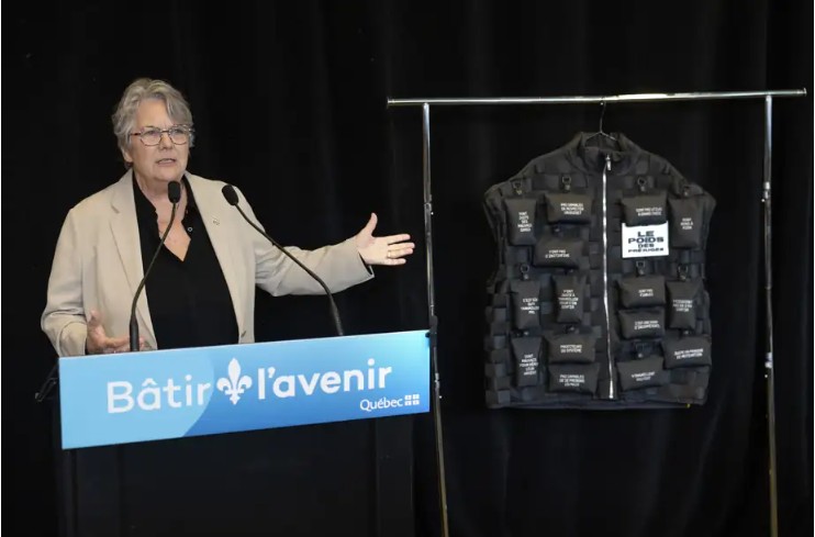 Woman in a beige blazer speaks at a podium labeled 'Bâtir l'avenir Québec' with a black tactical vest on a stand beside her, during a presentation.