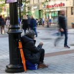 Person in a dark jacket sits on the pavement beside a lamppost, holding a coffee cup as pedestrians pass on a busy city street.