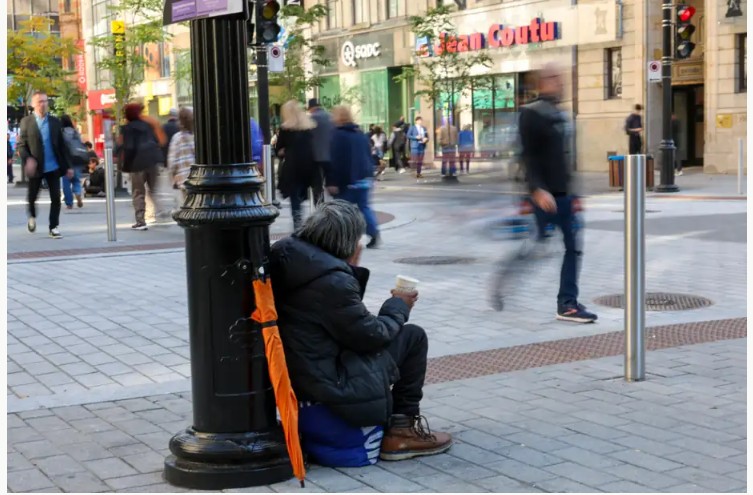Person in a dark jacket sits on the pavement beside a lamppost, holding a coffee cup as pedestrians pass on a busy city street.