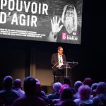 Male speaker at a clear podium giving a talk on a dark stage; a large screen behind him displays 'POUVOIR D’AGIR' in white letters.