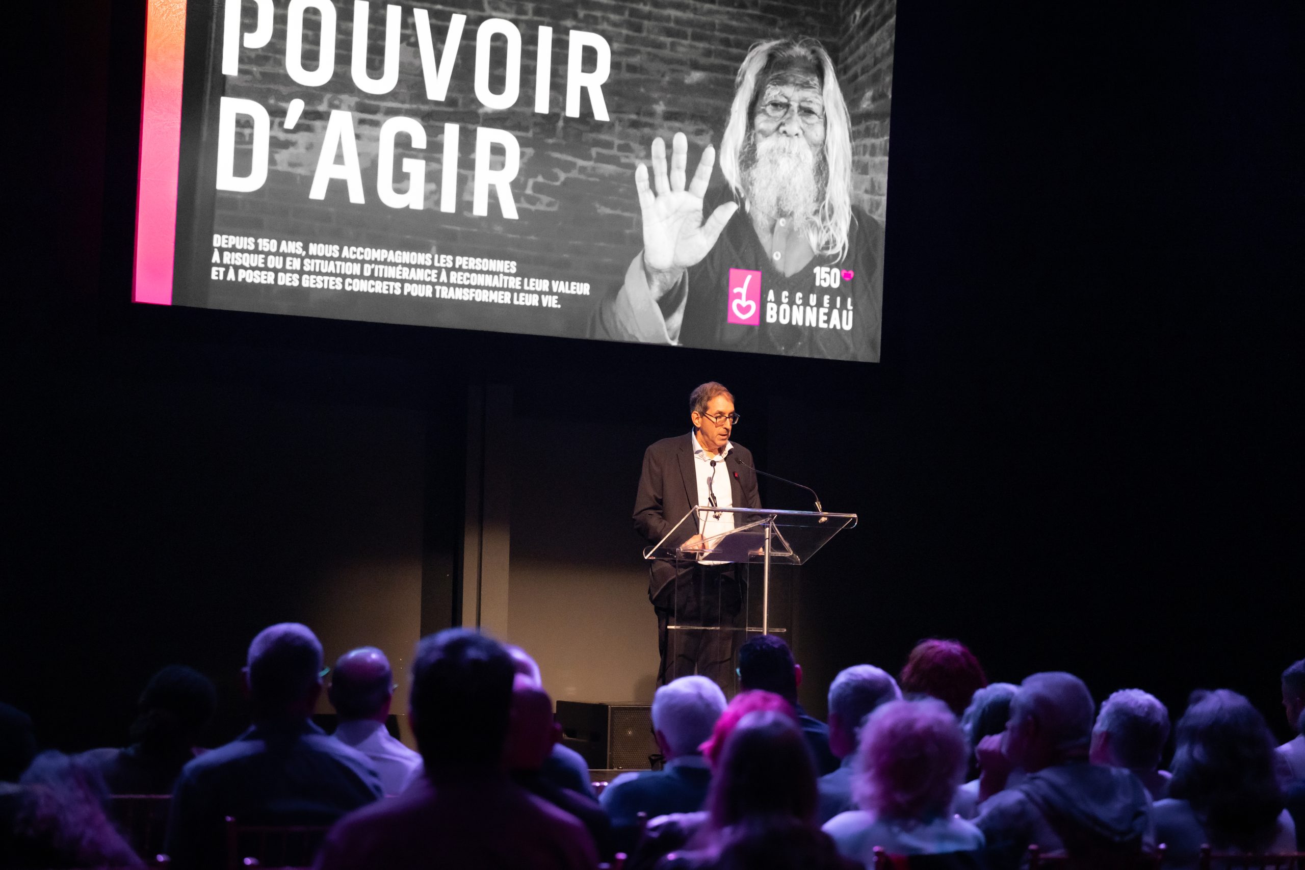 Male speaker at a clear podium giving a talk on a dark stage; a large screen behind him displays 'POUVOIR D’AGIR' in white letters.