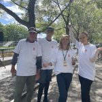 Four people in white shirts stand together outdoors under leafy trees on a sunny day, posing for a group photo.