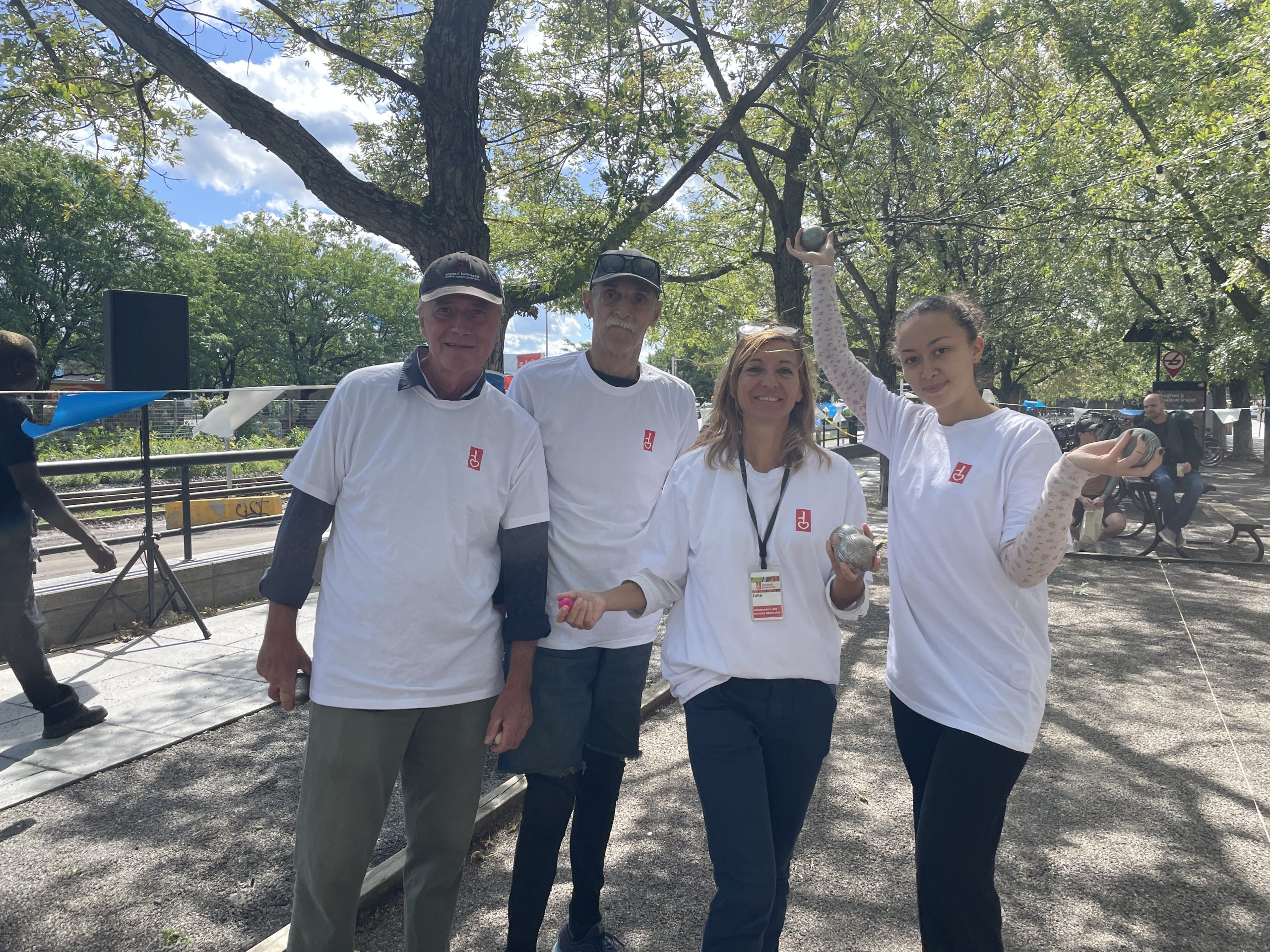Four people in white shirts stand together outdoors under leafy trees on a sunny day, posing for a group photo.